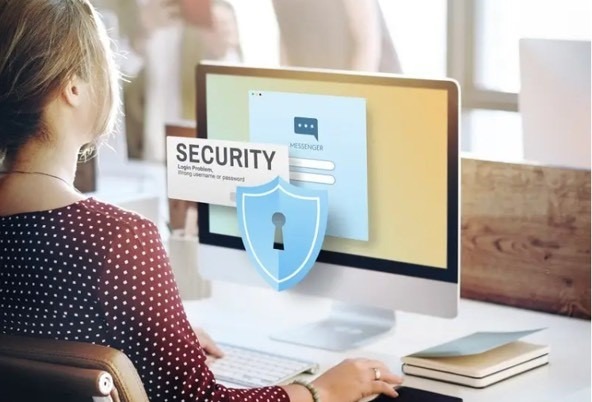 Mujer de espaldas. Pantalla de ordenador con imagen de un escudo con una cerradura en su centro y un cartel que dice "seguridad".