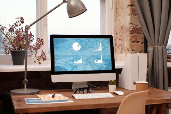 Escritorio con ordenador de mesa encendido en el que aparecen gráficos, además de eso en el escritorio hay un flexo, teclado, ratón y papeles